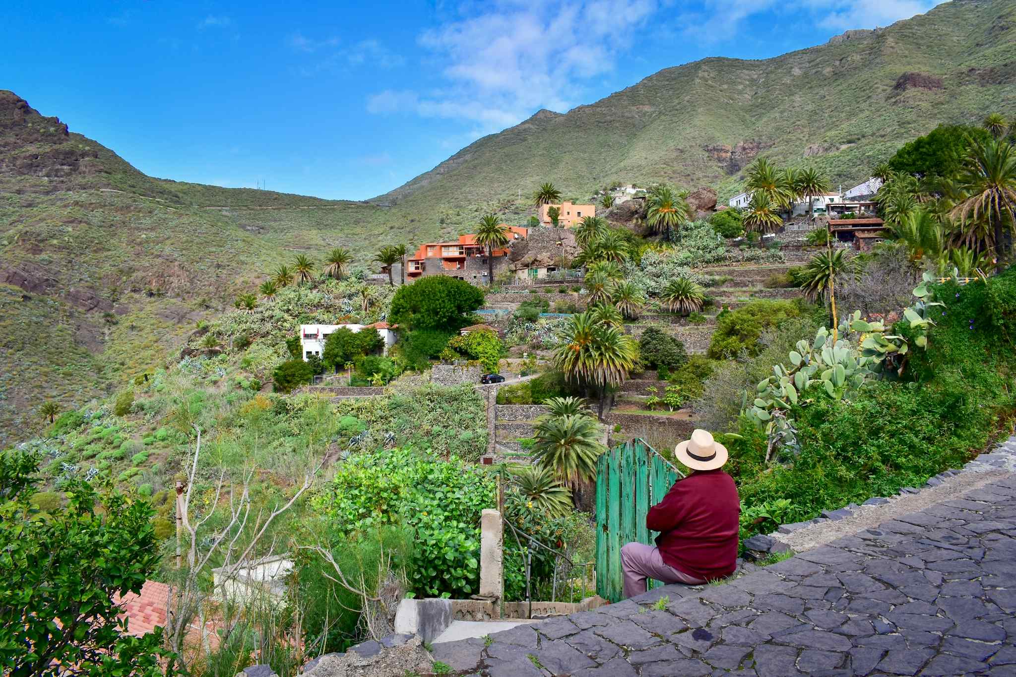 Man wearing a hat and red jumper sits on a wall and looks up at a small village in the hills
