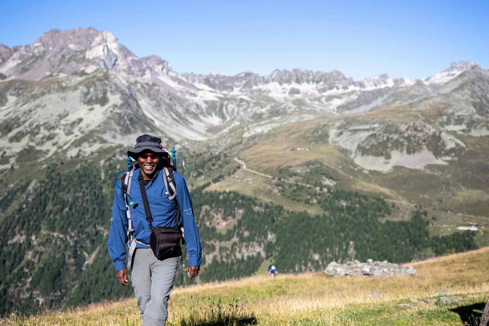 Man hiking up a grassy slope with high mountains in the background.