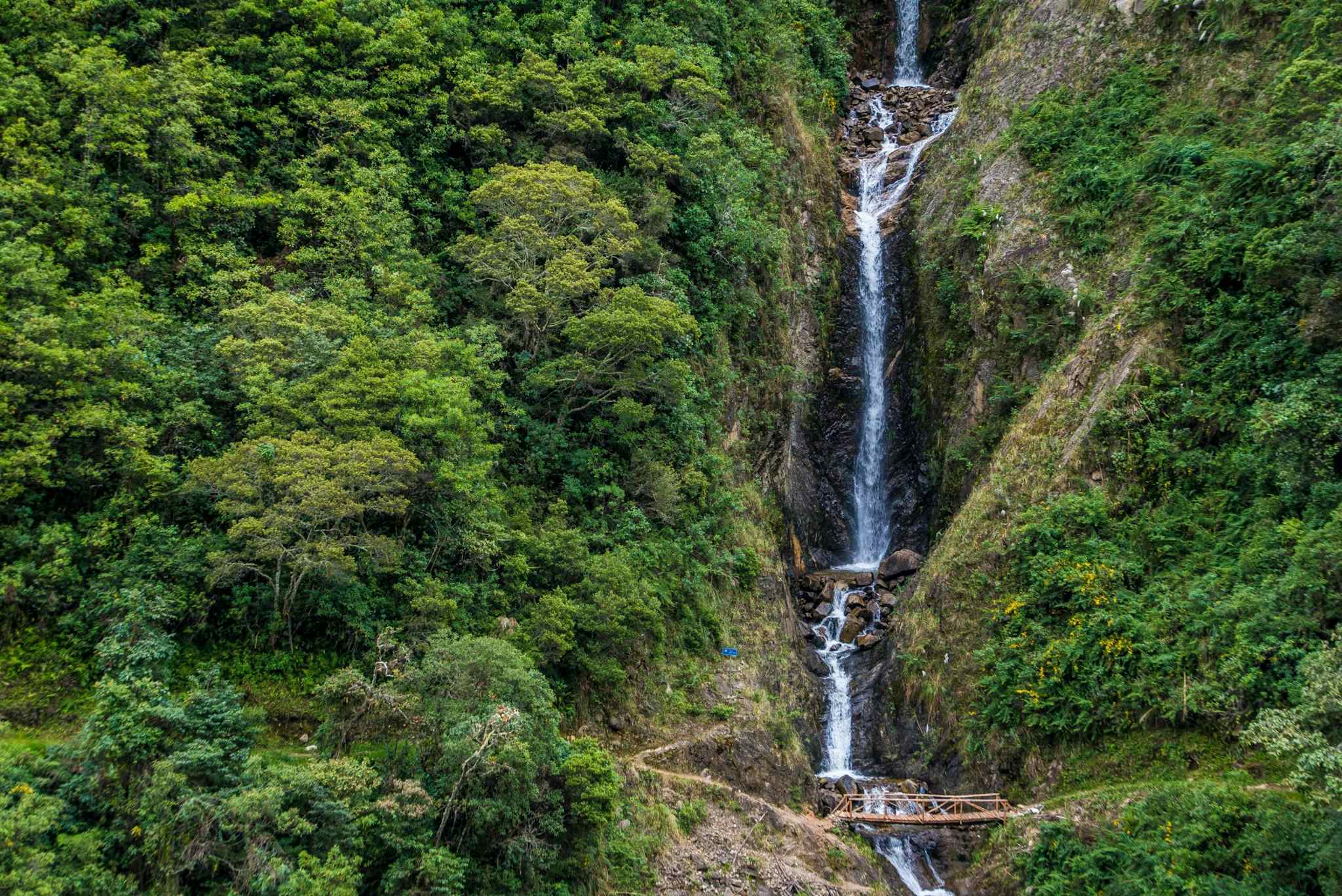Hikers crossing a wooden bridge below a tall waterfall surrounded by jungle in Peru.