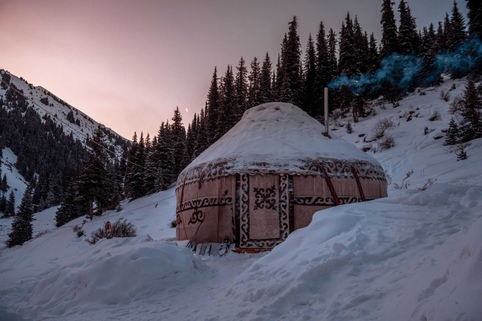 Yurt with a smoking chimney among a snowy landscape backed by forest and mountains in Kyrgyzstan.