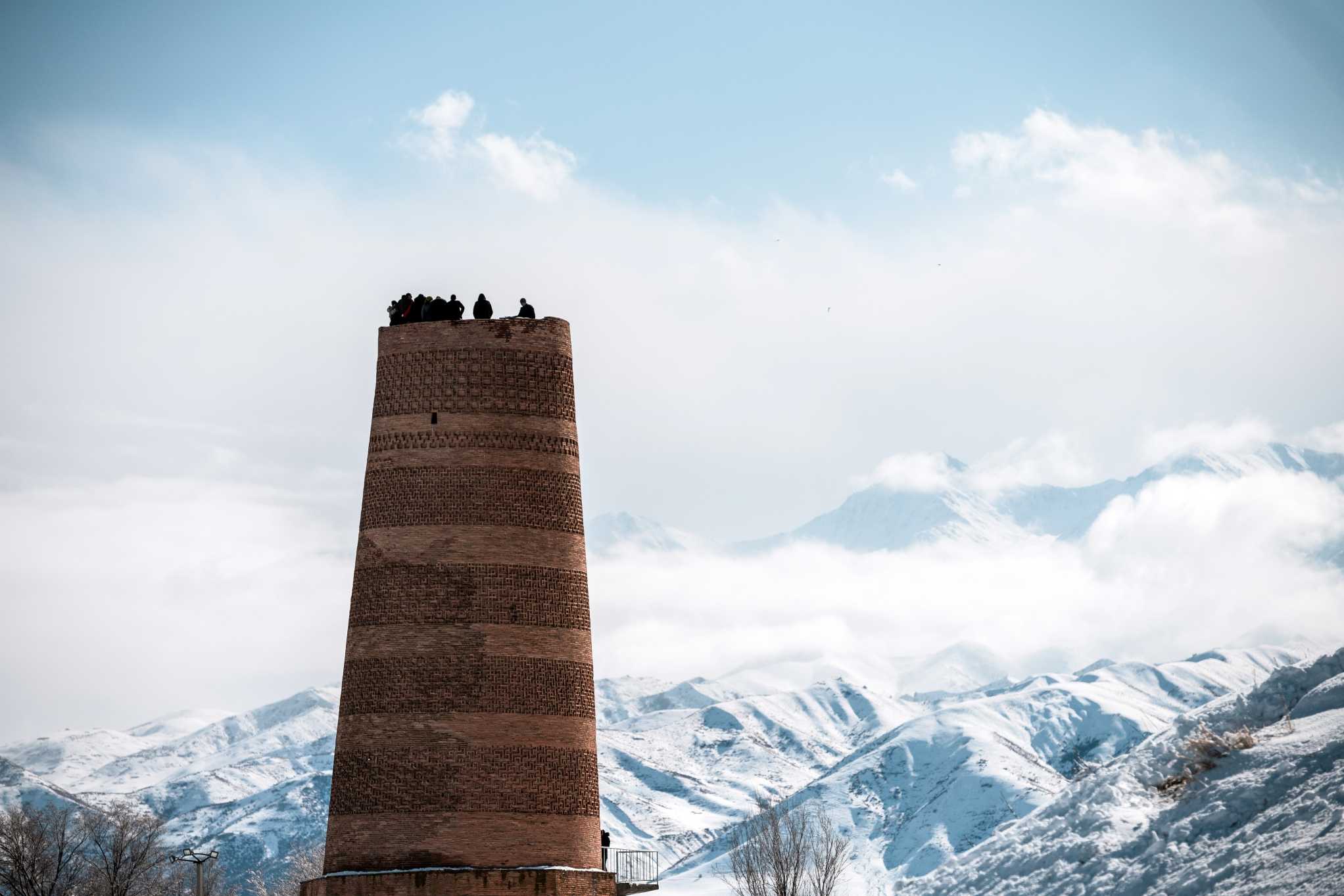 The Burana Tower in Kyrgyzstan with people on top and snowy mountains in the background.