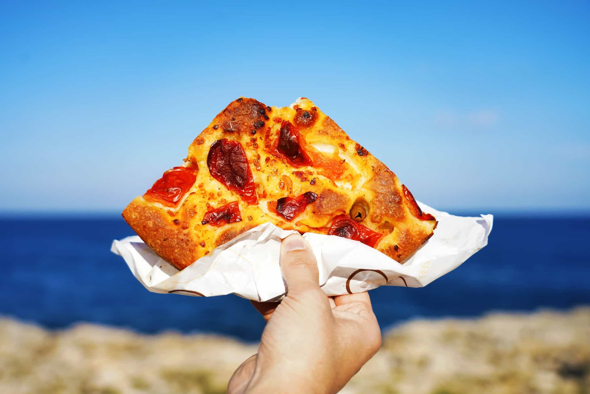 Hand holding a slice of focaccia with the sea in the background.