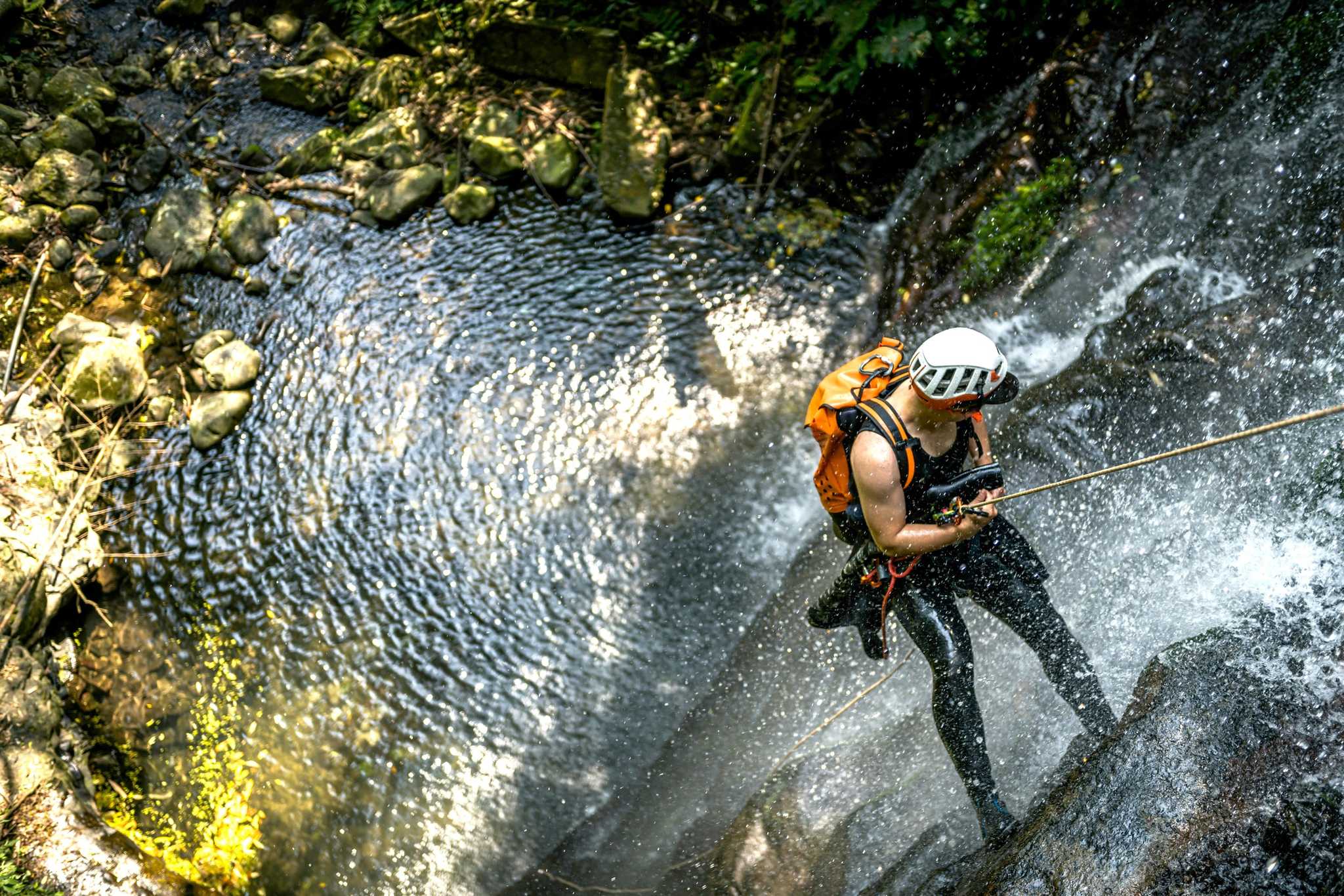 Man with an orange backpack rappelling down a waterfall into a pool of water below