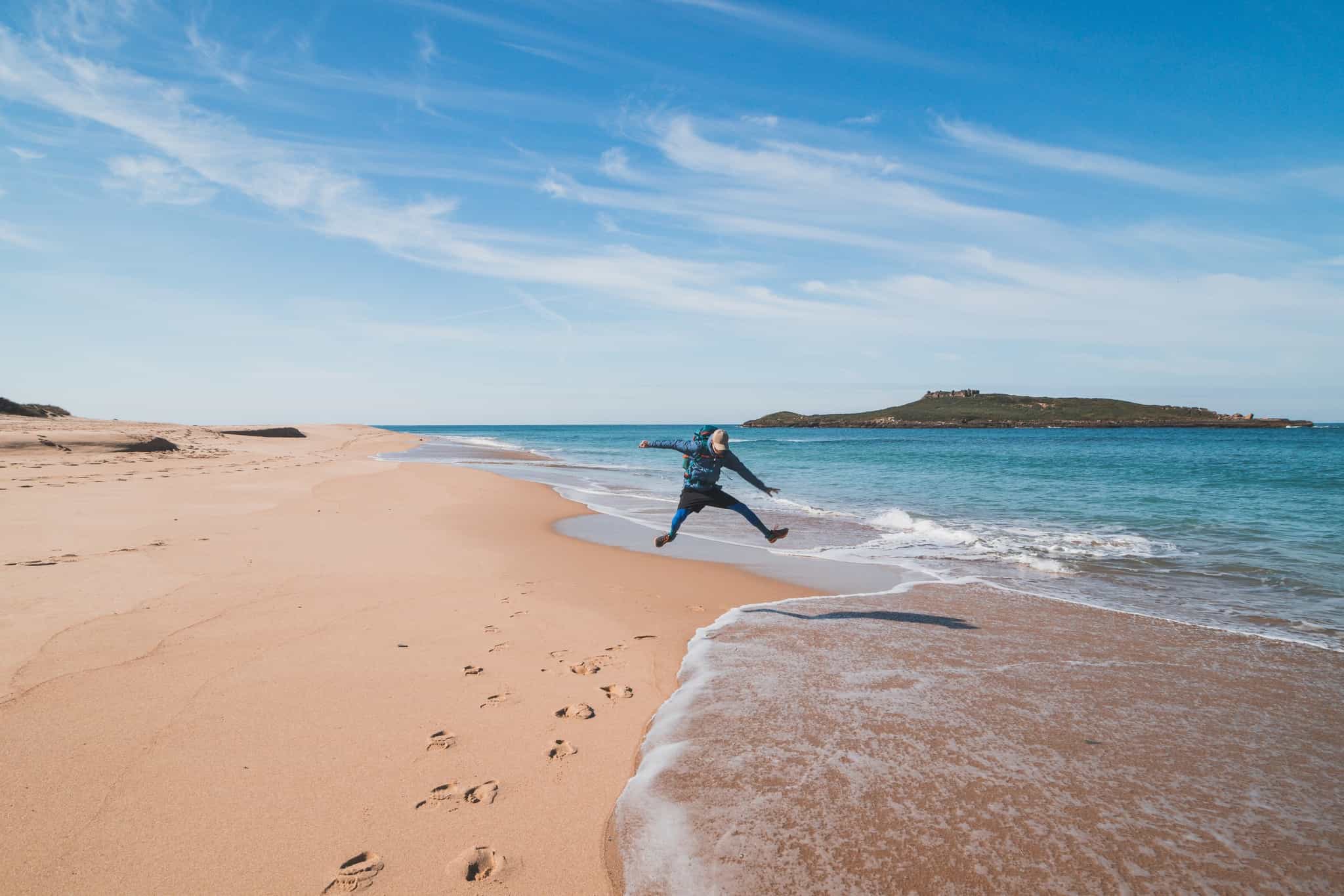 Hiker jumping the waves of the Atlantic Ocean on a sandy beach near Porto Covo, Portugal, by the Rota Vicentina.