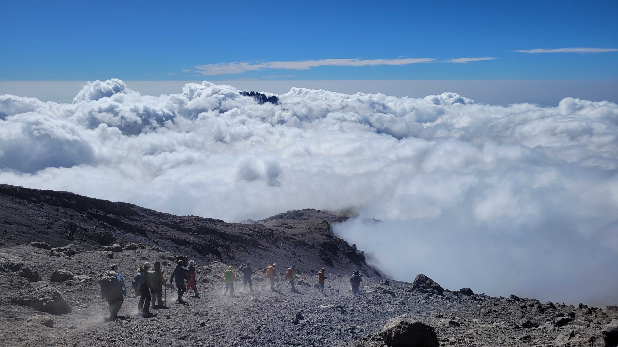 A line of trekkers following the dusty path down Kilimanjaro, above a blanket of clouds.