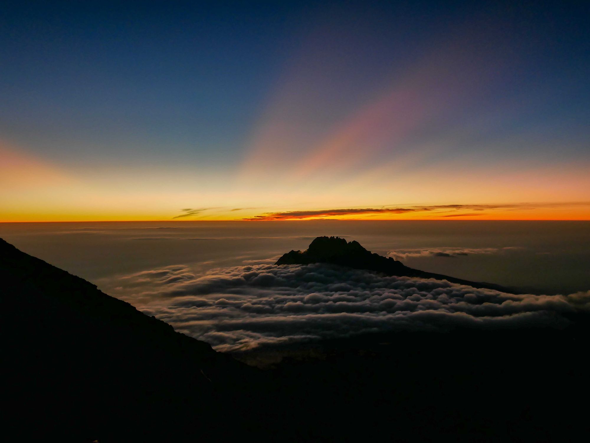 Rays of light emerging from the horizon, illumination Kilimanjaro's Mawenzi Peak, circled by cloud.