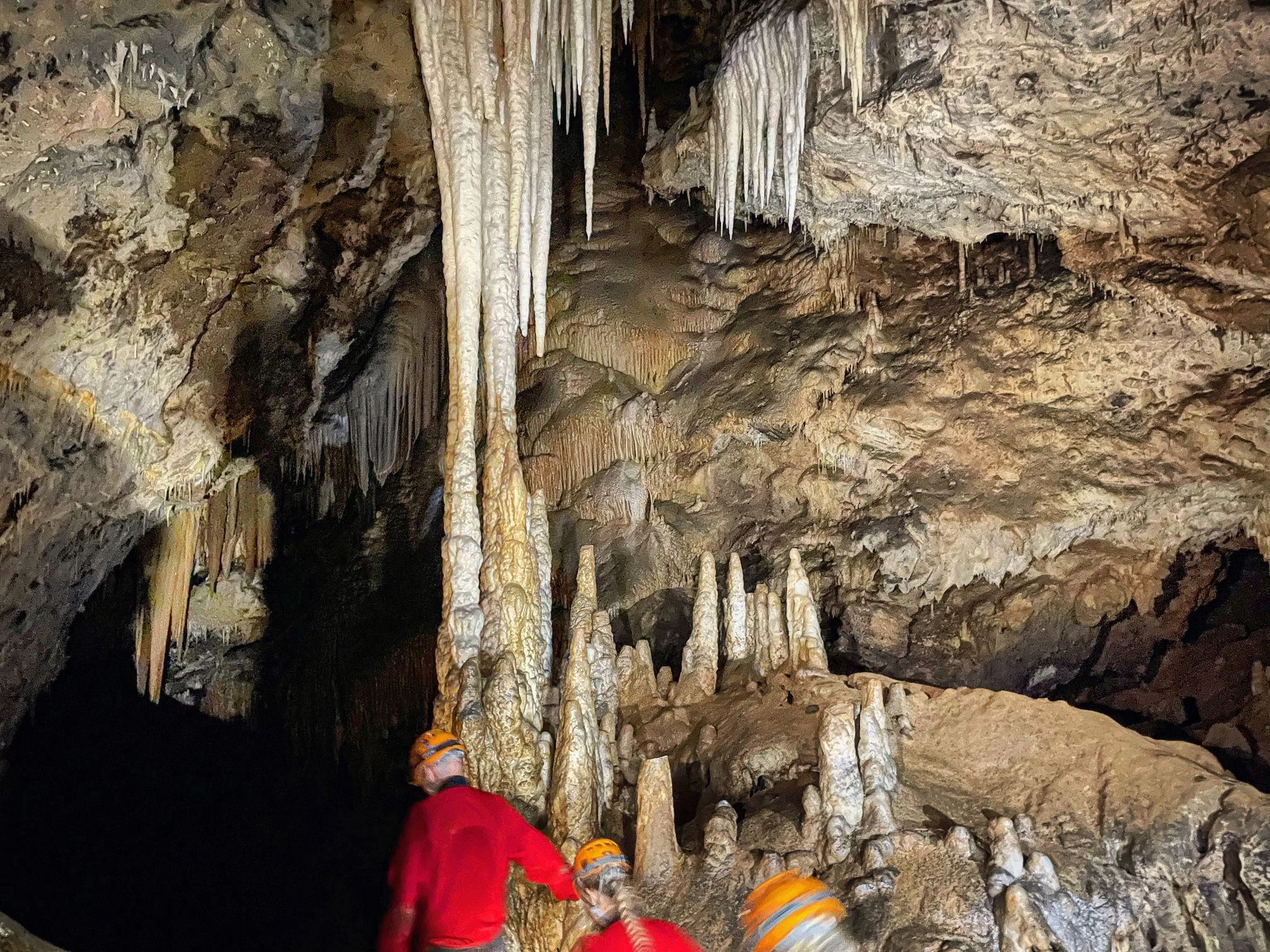 Three people heading into a dark cave, with many stalagmites and stalactites illuminated by torchlight.